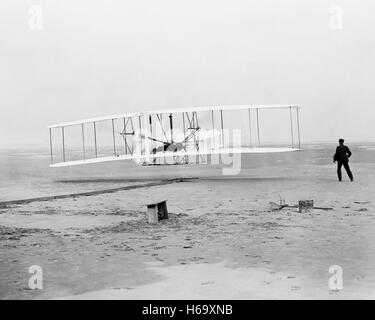 First flight, the Wright brothers, 120 feet in 12 seconds; Kitty Hawk, North Carolina.The photograph shows Orville Wright at the controls of the flying machine, lying prone on the lower wing with hips in the cradle which operated the wing-warping mechanism. Wilbur Wright running alongside to balance the machine, has just released his hold on the forward upright of the right wing. The starting rail, the wing-rest, a coil box, and other items needed for flight preparation are visible behind the machine. Photograph by John T. Daniels Stock Photo