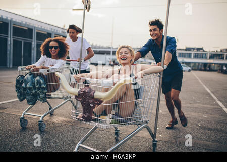 Young people racing with shopping trolleys on road. Multiracial group of friends racing with shopping cart. Stock Photo