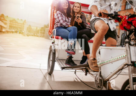 Young women sitting on tricycle and posing for selfie. Female friends enjoying tricycle ride on road and taking self portrait wi Stock Photo