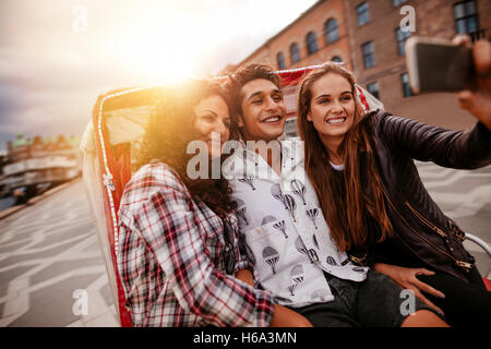 Teenage friends having fun on vacation, traveling on tricycle and taking selfie with mobile phone. Young man and women riding on Stock Photo