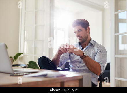 Shot of young man sitting at table looking at laptop and thinking. Thoughtful businessman working at home office. Stock Photo