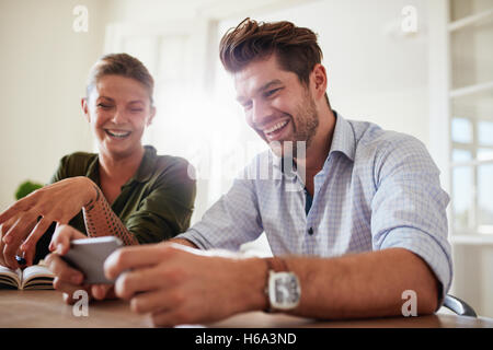 Shot of young couple sitting at table using cellphone at home and smiling. Man and woman smiling with mobile phone. Stock Photo