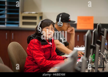 A middle-aged Asian female Santa Ana, CA, community college student works at a learning skills center computer monitor. Note headphones and Hispanic student in background. Stock Photo
