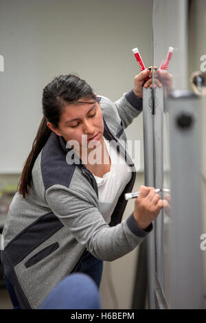 A Hispanic teacher writes on a white board with a felt tip marker in the learning center of a Santa Ana, CA, community college. Note reflection. Stock Photo
