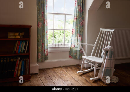 A white, empty rocking chair on a solid wood floor by a window in a room with a throw over it's arm. Stock Photo