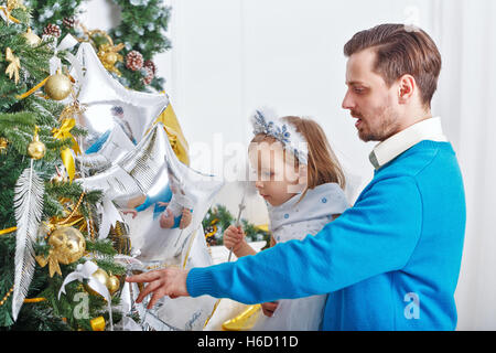 Daughter and father decorate the Christmas tree. Father supports daughter and little cute girl hangs on the Christmas tree toys. Stock Photo
