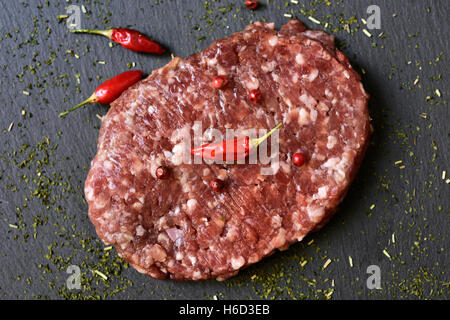 closeup of raw burger spiced with red peppercorns, chili peppers and herbs on a slate surface