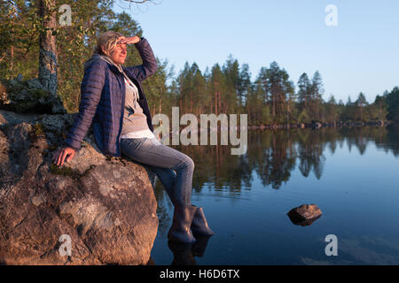 Beautiful tourist woman is sitting on the stone near of the sunset lake and looking inti the distance Stock Photo