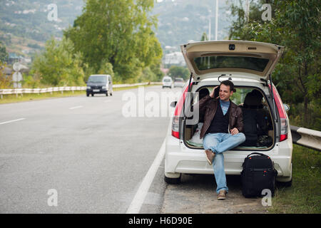 Calling friends. Happy young man talking mobile phone and sitting on a car trunk Stock Photo