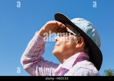 A senior woman wearing a wide brimmed sunhat and dark glasses in sunshine shades her eyes to look far up at the sun in summer. England UK Britain Stock Photo