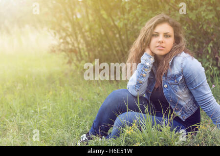 Portrait of cool long haired brunette teenage girl sitting outdoors in sun light. Giirl is looking at the camera. Stock Photo