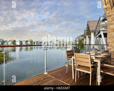 A set dining table & chairs on decking overlooking a lake surrounded by holiday homes on a lovely evening in the Cotswolds, UK Stock Photo