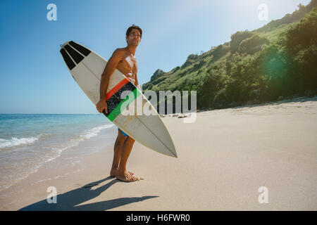 Full length shot of young man with surf board on beach. Male surfer holding surfboard on the sea shore and looking away. Stock Photo