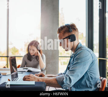 Shot of young man with headphones using laptop at college library.  Young people sitting at table working on school assignment Stock Photo