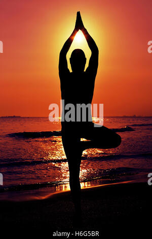 the silhouette of a young yogi man practicing the tree pose in front of the sea in backlight Stock Photo