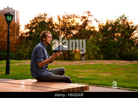 Guy sitting on a bench in the park reading book Stock Photo