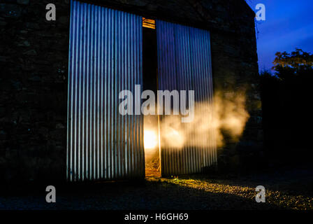 Light shines through smoke coming out from half open doors of an old Irish stone barn at night Stock Photo