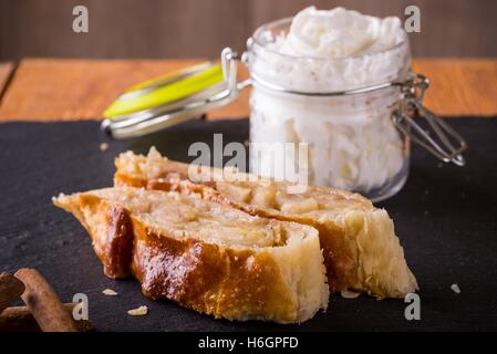 Horizontal photo of apple strudel pie on black slate stone and wooden board. Few pieces of cinnamon next to sweets. White whippe Stock Photo