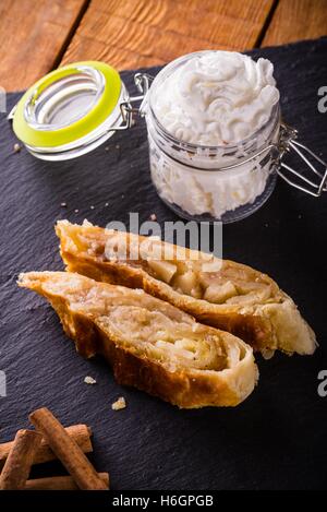 Vertical photo of apple strudel pie on black slate stone and wooden board. Few pieces of cinnamon next to sweets. White whipped  Stock Photo