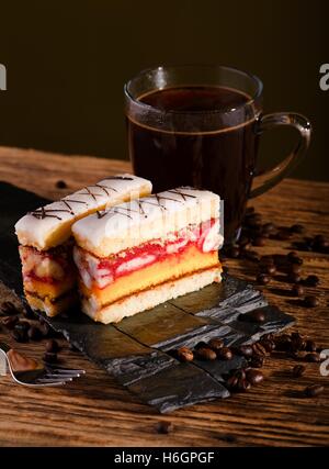 Vertical photo of two slices of sweet desert with color layers on sugar on the top placed on piece of black stone. Cup of coffee Stock Photo