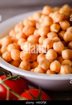 Vertical photo of white bowl with wet yellow chickpeas on wooden board. few tomatoes are in front of legumes. Stock Photo