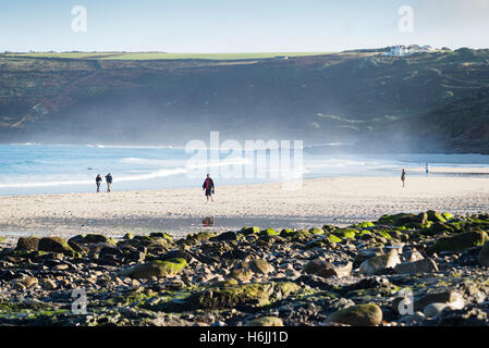 People walking over the sandy beach in the spray of the Atlantic ocean surf at low tide after sunrise at Sennen Cove, west coast of Cornwall,UK Stock Photo