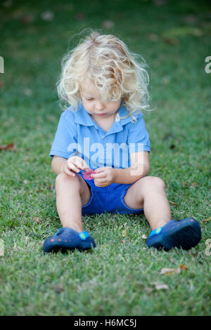 Beautiful boy three year old with long blond hair sitting on the grass Stock Photo
