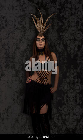 Tanned female model in amazonian superhero style outfit with feather headdress, black damask backdrop Stock Photo