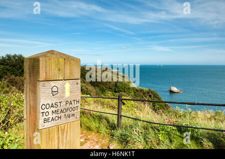 Coast Path 'To Meadfoot Beach 1/4 mile' sign post next to footpath with sea and Shag Rock in background on clear summer's day. Stock Photo