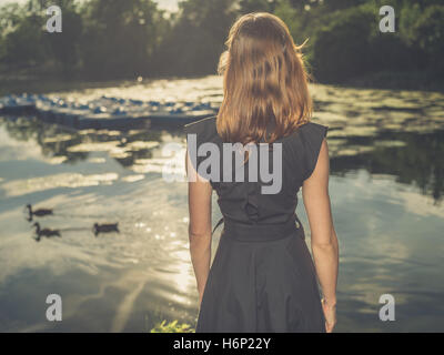 Vintage filtered shot of an elegant young woman standing by a lake in a park at sunset Stock Photo