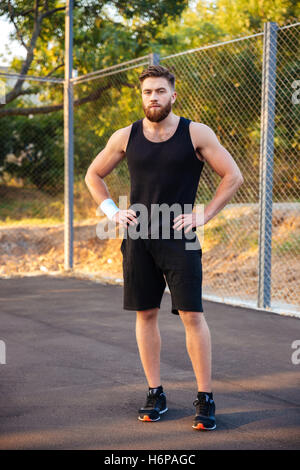 Concentrated young bearded man in sportswear standing with hands on hips outdoors Stock Photo
