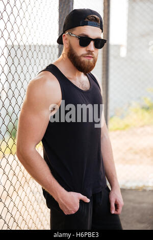 Portrait of handsome bearded man leaning on metal urban fence wearing sunglasses and cap Stock Photo