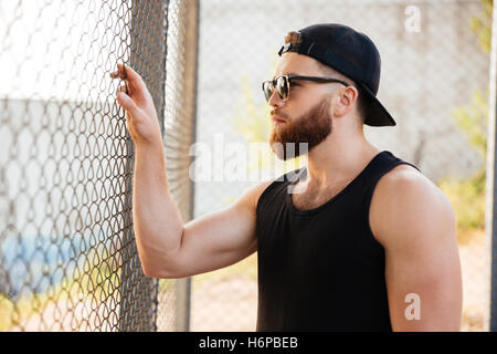 Close up portrait of young bearded man looking through metal urban fence wearing sunglasses and cap outdoors Stock Photo