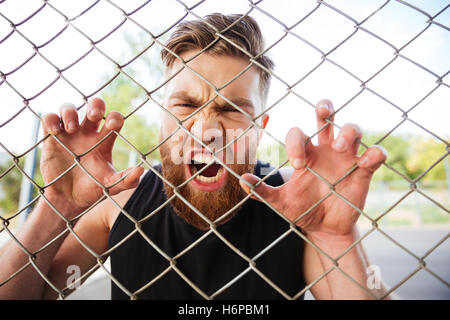 Close up portrait of a crazy bearded man shouting with his hands on metal fence outdoors Stock Photo