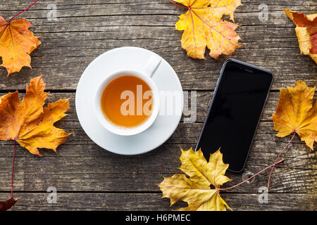 Cup of tea and smartphone on wooden table with autumn leaves. Top view. Stock Photo