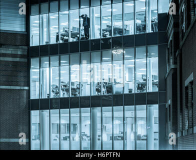 Man alone in office looking out of window at night: energy saving, working from home workplace stress, mental health, loneliness...concept. Stock Photo