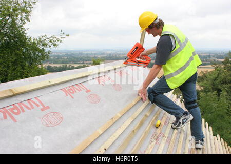 A single roofer nails timber battens to the roof of a timber house on an Oxfordshire hillside. Shows wood and fields beyond. Stock Photo