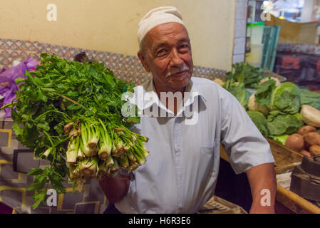 Smiling male Moroccan stallholder wearing a Kufi hat, holding bunches of beldi (rural) celery also known as Wild celery (Apium graveolens) at a local market in Marrakech, Morocco. Stock Photo