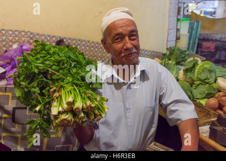 Smiling male Moroccan stallholder wearing a Kufi hat, holding bunches of beldi (rural) celery also known as Wild celery (Apium graveolens) at a local market in Marrakech, Morocco. Stock Photo