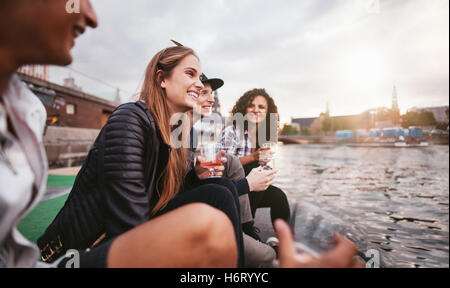 Shot of group of happy young people relaxing by the lake and having drinks. Teenage friends sitting on jetty and smiling. Stock Photo