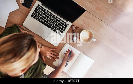 Top view shot of woman sitting at table with laptop and coffee writing on notebook. Female making to do list on diary. Stock Photo