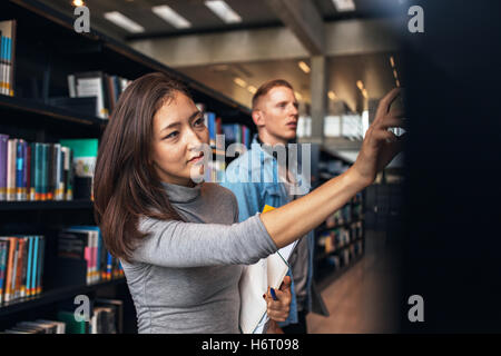 Shot of young female student selecting book from library shelf with man in background. university students taking book from shel Stock Photo