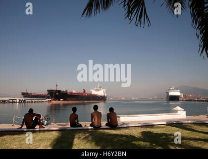 Young Men sitting on the dock side in the shade, watching ships coming in to the harbour, Manzanillo, Mexico Stock Photo
