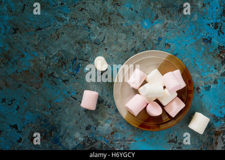 White and pink marshmallows in a ceramic bowl on blue concrete background. View from above. Stock Photo