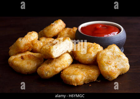 Pile of golden deep-fried battered chicken nuggets with empty rustic bowl on dark wood. One cut with meat showing. Stock Photo