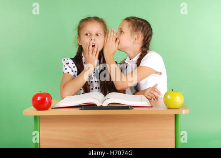 Two school girl sharing secrets  sitting at a desk from book isolated on green Stock Photo