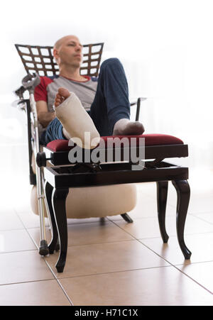 Young man with a broken ankle and a white cast on his leg, sitting on a couch, with crutches nearby, looking sad and frustrated Stock Photo