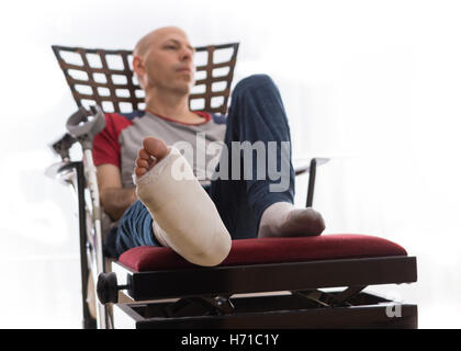 Young man with a broken ankle and a white cast on his leg, sitting on a couch, with crutches nearby, looking sad and frustrated Stock Photo