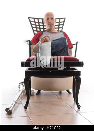 Young man with a broken ankle and a white cast on his leg, sitting on a couch, with crutches nearby, looking bored Stock Photo