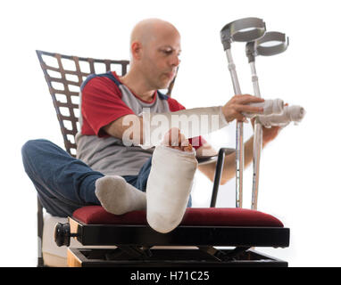 Young man after a bad accident with broken ankle and wrist and white plaster and fiberglass casts on his leg and arm, putting do Stock Photo